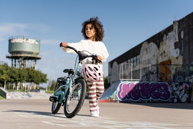 Ett litet barn med lockigt hår, iklädd vit skjorta och randiga byxor, går bredvid en blå PUKY Barncykel - 16 Tum - 4 år - 1 Gear - Puky Youke 16-cykel medan han håller i en rosa hjälm i en stadsdel dekorerad med graffiti. Barnet tittar åt sidan och ett vattentorn syns i bakgrunden.