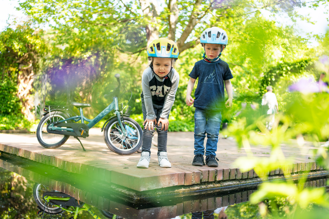 Två barn som bär hjälmar står på en tegelplattform bredvid en reflekterande vattenkropp. Ett barn, fascinerat av sin PUKY Barncykel - 16 Tum - 4 år - 1 Gear - Puky Youke 16:s lättviktskomponenter, hukar sig för att titta på vattnet medan det andra står i närheten. En PUKY barncykel står parkerad i närheten. Den frodiga gröna bakgrunden föreslår en park eller naturmiljö.