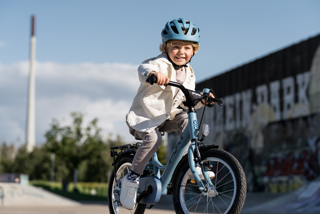 Ett litet barn med ljusblå hjälm och vit jacka åker PUKY Barncykel - 16 Tum - 4 år - 1 Gear - Youke 16 i en park. Barnet ler brett och verkar njuta av åkturen. I bakgrunden finns en lång vägg med graffiti och en hög struktur i fjärran.