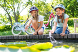 Två unga flickor i hjälmar leker vid en damm och stänker vatten med händerna. Parkerade bakom dem står deras PUKY Juniorcykel - 20 Tum - 6 år - 3 Gear - Dynamo Lampa - Puky Skyride Classic cyklar. Inställningen verkar vara en park med grönskande träd, vilket tyder på ett soligt äventyr utomhus. Båda tjejerna ler och njuter av stunden.